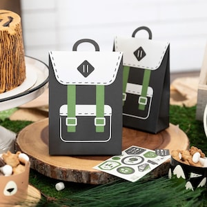 May include: Two black and white paper favor bags shaped like backpacks with green straps. The bags are sitting on a wooden slice. There are stickers with a camping theme on the table.