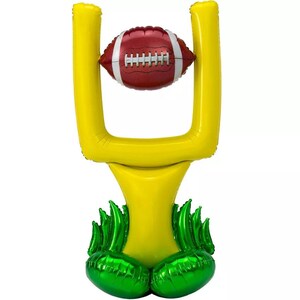 May include: A yellow inflatable football goal with green grass-like accents at the base. A brown football is positioned in the goal.