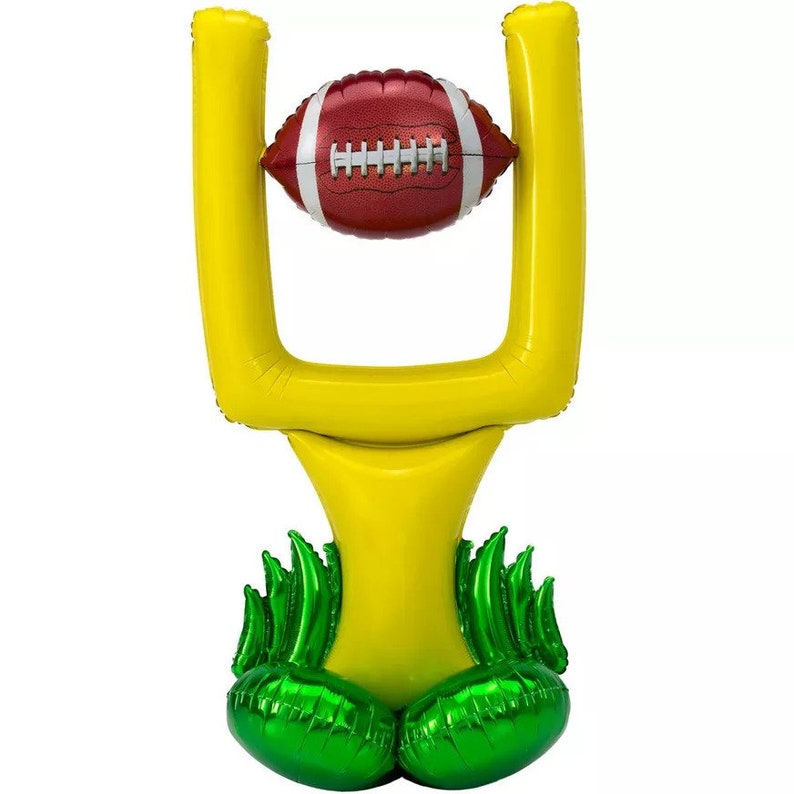 May include: A yellow inflatable football goal with green grass-like accents at the base. A brown football is positioned in the goal.