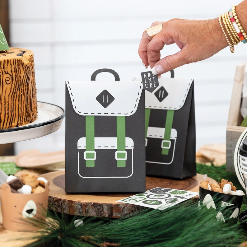 May include: Two black and white paper bags with a green backpack design. The bags have a white label that says "Adventure" on them. The bags are sitting on a wooden surface with a brown cake and a cup of snacks in the background.
