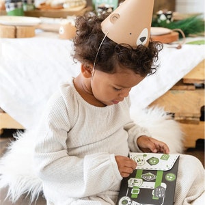 May include: A child wearing a brown bear party hat is playing with a black and green paper backpack with stickers. The backpack has the words "WANDER", "EXPLORE", "ADVENTURE", and "LOVE" on it.