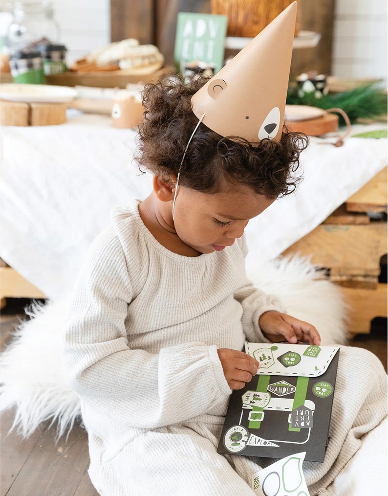 May include: A child wearing a brown bear party hat is playing with a black and green paper backpack with stickers. The backpack has the words "WANDER", "EXPLORE", "ADVENTURE", and "LOVE" on it.