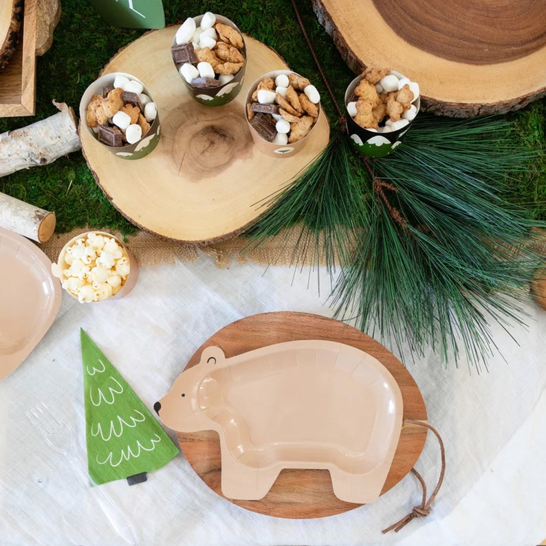 May include: A brown wooden plate with a beige bear-shaped paper plate on top. The bear plate has a brown outline and a white interior. The plate is on a white tablecloth with a green pine branch and a green tree-shaped napkin.