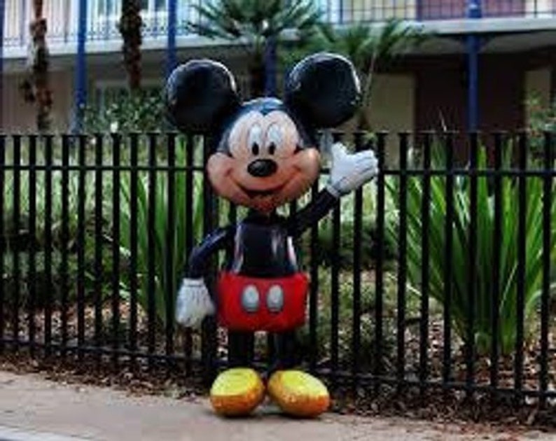 Puede incluir: Un gran globo de Mickey Mouse negro y rojo con una gran sonrisa y guantes blancos. El globo est&aacute; de pie frente a una valla de metal negro.