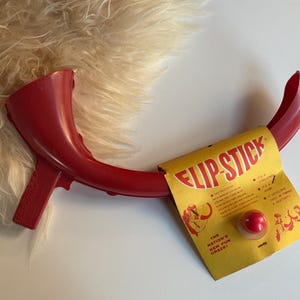 May include: A red plastic Flip-Stick toy with a yellow and red label. The label reads "Flip-Stick" and includes illustrations and text. The toy has a curved design with a handle and a red ball.