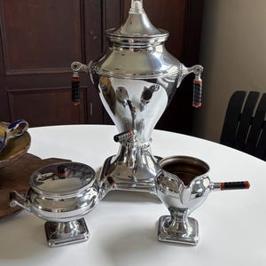 May include: A polished silver-tone metal coffee service set consisting of a tall urn with a glass finial, a lidded sugar bowl, and a creamer. Each piece features square bases and black ribbed handles with orange accents, creating a cohesive metallic aesthetic.