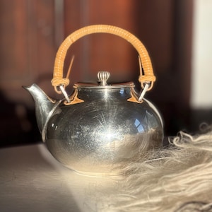 May include: A silver teapot with a rounded body and a curved spout. The teapot has a woven handle and a small, decorative knob on the lid. The teapot is sitting on a white surface.
