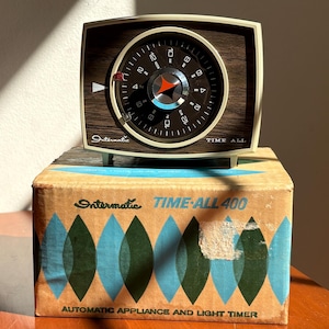 May include: An Intermatic Time-All 400 automatic appliance and light timer with a wood-grain face and a circular clock dial. The device sits atop its original cardboard box, which features a repeating blue and green leaf-like pattern.
