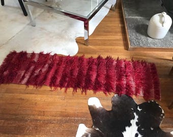 May include: A rectangular, deep red rug with a shaggy texture. The rug is placed on a hardwood floor, with a cowhide rug and a glass-topped table in the background. The rug's texture appears soft and plush.