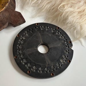 May include: A black circular mold with a central hole and a ring of small, raised, decorative designs around the edge.