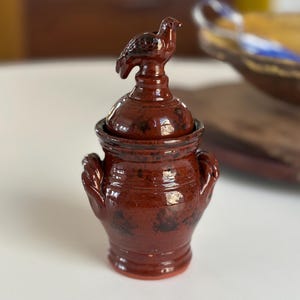 May include: A small, brown ceramic jar with a bird-shaped lid. The jar has two handles and a glazed finish.
