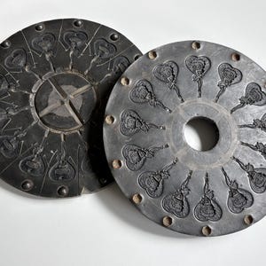 Peut inclure: Deux moules circulaires en métal gris foncé présentant de multiples cavités en creux disposées en motif radial. Chaque cavité contient un motif en relief détaillé d'un personnage de dessin animé dans un cœur. Les moules ont une ouverture centrale et des encoches circulaires sur le bord extérieur.
