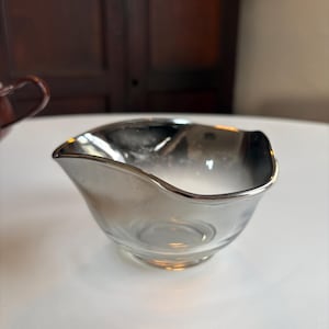 May include: A clear glass gravy boat with a silver-colored rim. The boat has a curved, asymmetrical shape with a spout on one side. The interior is transparent, and the boat sits on a small, round base.