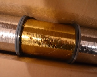 May include: Two spools of metallic thread, one gold and one silver, are wound tightly together. The spools are in a cardboard box.