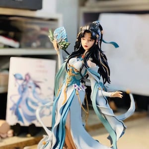 K&ouml;nnte beinhalten: Detaillierte Figur einer Frau in einem flie&szlig;enden, hellblauen und wei&szlig;en Kleid, die einen F&auml;cher h&auml;lt. Die Figur hat langes schwarzes Haar und steht auf einem rosa-pfirsichfarbenen Sockel. Das Kleid hat goldene Akzente und florale Details.
