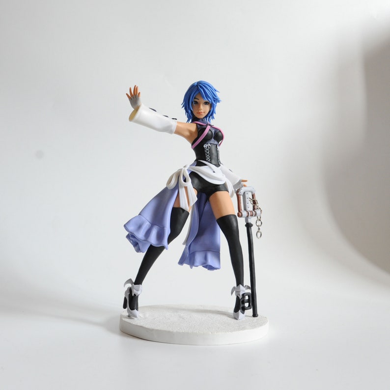 May include: A figurine of a character with blue hair, wearing a black and white outfit with a purple skirt. The figure is holding a staff with a clock-like design. The figurine is standing on a white base.