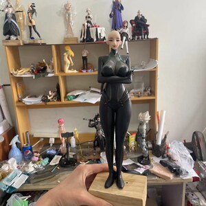 May include: A black figurine of a woman with a bald head and a futuristic outfit, standing on a wooden block. The figurine has her arms crossed and is surrounded by other figurines on a shelf.