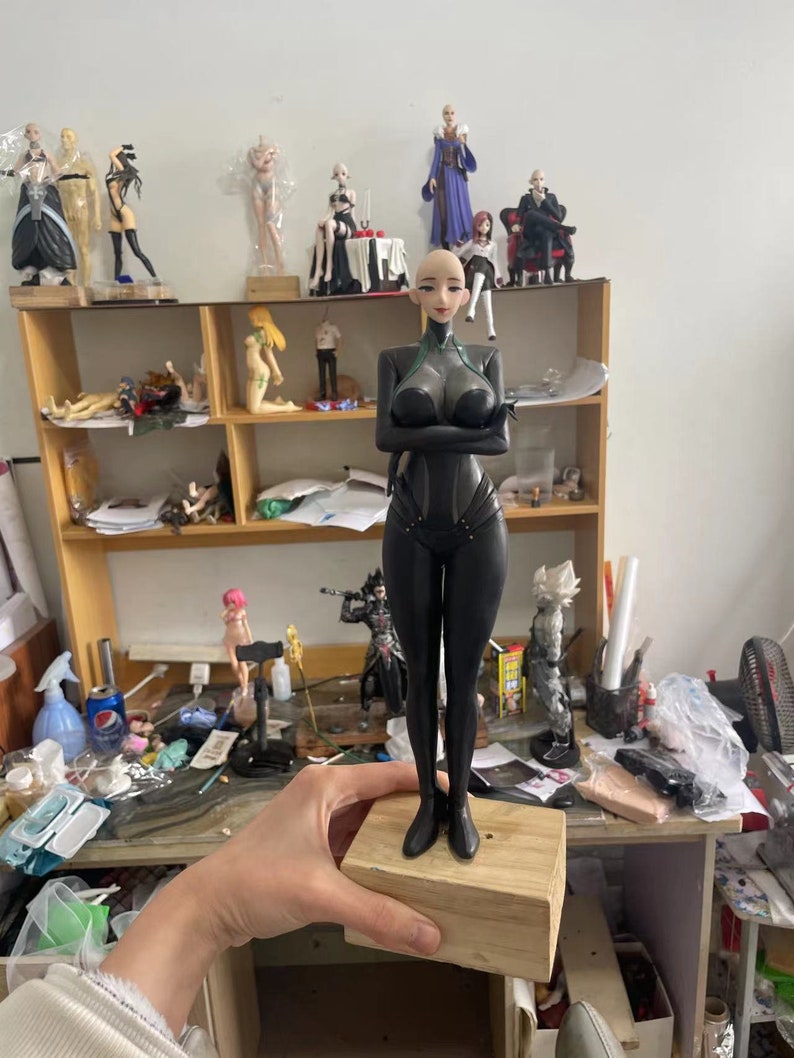 May include: A black figurine of a woman with a bald head and a futuristic outfit, standing on a wooden block. The figurine has her arms crossed and is surrounded by other figurines on a shelf.