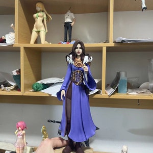 May include: A detailed figurine of a woman in a long, flowing purple dress with brown accents and a decorative bodice. The figure has long brown hair and is holding a small object. Other figurines are visible on shelves in the background.