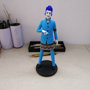 K&ouml;nnte beinhalten: Handgefertigte Figur einer Figur mit blauen Haaren und einem blauen Anzug. Die Figur steht auf einem schwarzen, kreisf&ouml;rmigen Sockel. Das Outfit der Figur umfasst ein braunes, rockartiges Detail und schwarze Schuhe. Die Figur hat einen blassen Teint und ein stilisiertes Aussehen.