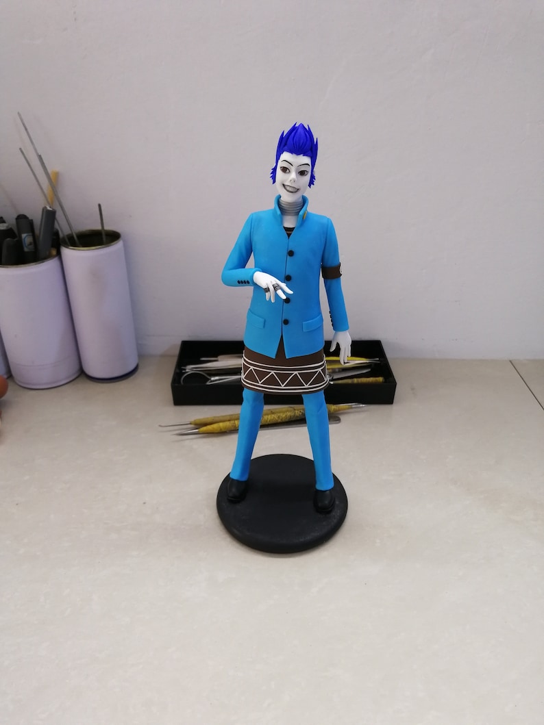 K&ouml;nnte beinhalten: Handgefertigte Figur einer Figur mit blauen Haaren und einem blauen Anzug. Die Figur steht auf einem schwarzen, kreisf&ouml;rmigen Sockel. Das Outfit der Figur umfasst ein braunes, rockartiges Detail und schwarze Schuhe. Die Figur hat einen blassen Teint und ein stilisiertes Aussehen.