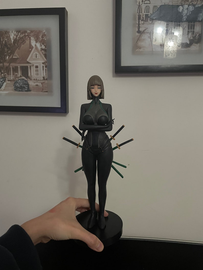 May include: A black figurine of a woman in a form-fitting outfit, with multiple swords attached. The figure has short brown hair and is standing on a black circular base. The swords have black blades and green handles.