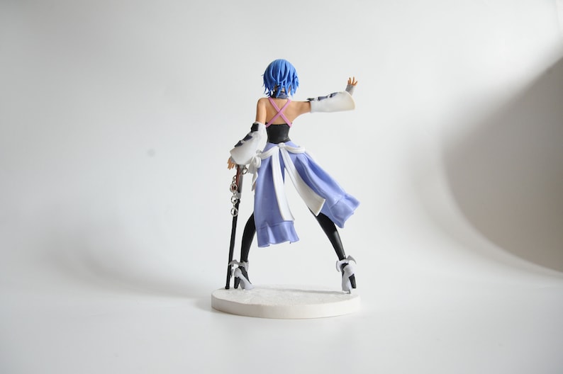 May include: A figurine of a character with blue hair, wearing a black top, a light purple skirt, and white accents. The figure is holding a staff and is standing on a white base. The figurine is approximately 7 inches tall.