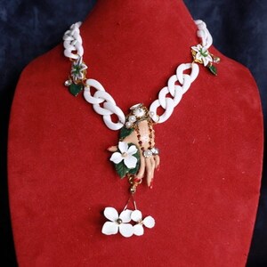 May include: White chunky chain necklace with a hand holding white flowers and a dangling cluster of white flowers. The hand is decorated with red and gold accents.