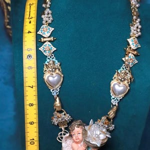 May include: Ornate gold-tone necklace with pearl accents, turquoise and white enamel details, and a central cherub figurine holding a flower. The necklace is approximately 10 inches long.