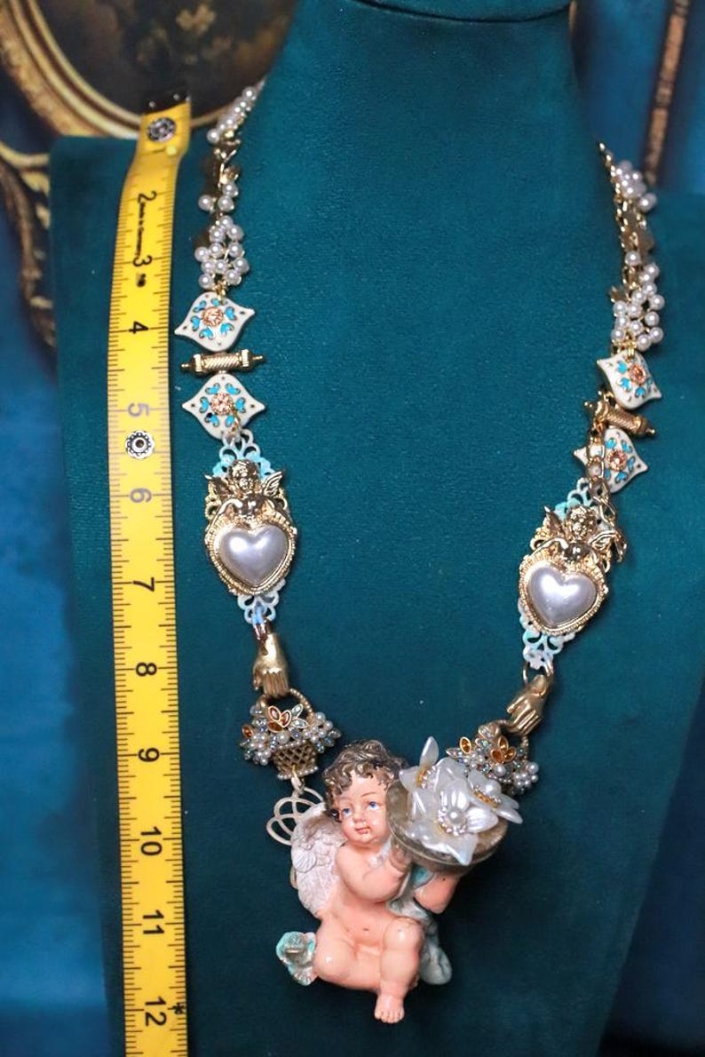 May include: Ornate gold-tone necklace with pearl accents, turquoise and white enamel details, and a central cherub figurine holding a flower. The necklace is approximately 10 inches long.