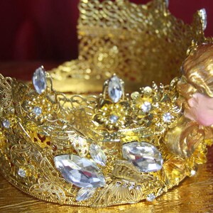 May include: A gold crown with intricate details and clear rhinestones. The crown features a small angel figurine with gold wings and a pink face.