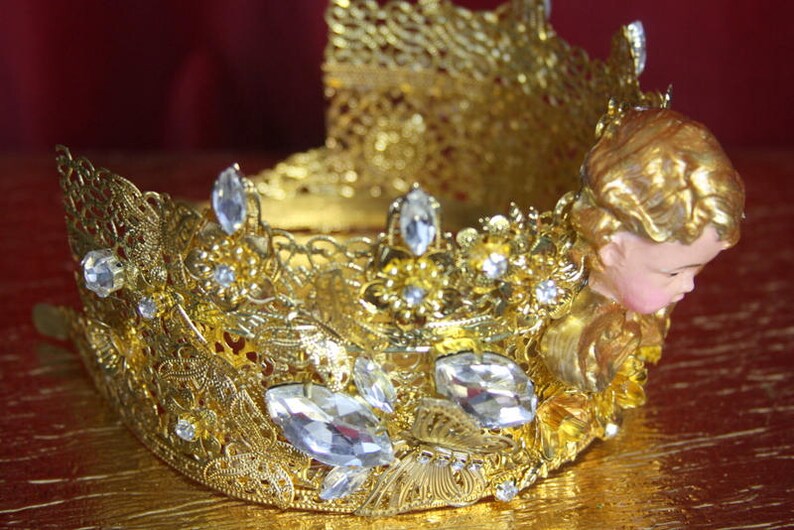 May include: A gold crown with intricate details and clear rhinestones. The crown features a small angel figurine with gold wings and a pink face.