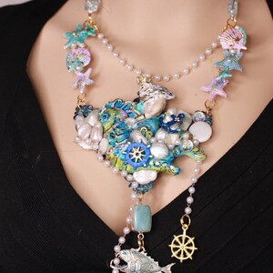 May include: A multi-layered necklace with a large, blue, green, and white seashell pendant. The pendant is decorated with pearls, starfish, and a small anchor. The necklace also features a smaller, blue fish pendant with a gold anchor charm.