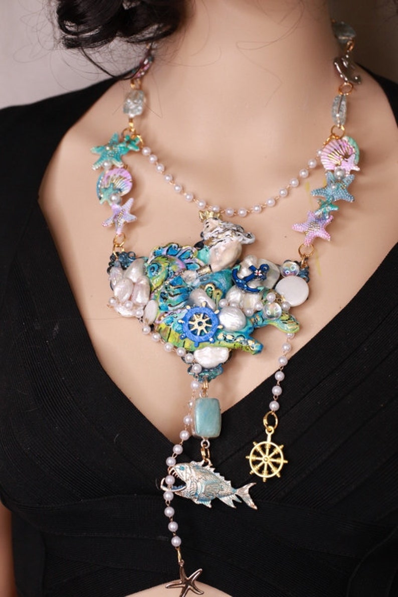 May include: A multi-layered necklace with a large, blue, green, and white seashell pendant. The pendant is decorated with pearls, starfish, and a small anchor. The necklace also features a smaller, blue fish pendant with a gold anchor charm.