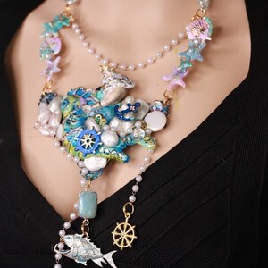 May include: A multi-layered necklace with a large, colorful, and textured centerpiece featuring a variety of sea-themed charms, including seashells, starfish, and a ship's wheel. The necklace is made of pearls, beads, and metal.