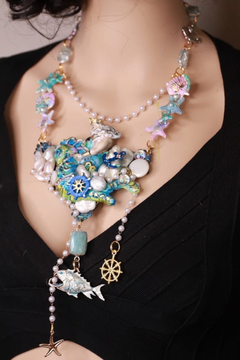 May include: A multi-layered necklace with a large, colorful, and textured centerpiece featuring a variety of sea-themed charms, including seashells, starfish, and a ship's wheel. The necklace is made of pearls, beads, and metal.