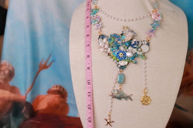 May include: A multi-strand necklace with a large, colorful pendant featuring seashells, starfish, and a fish. The pendant is made of various materials, including beads, metal, and resin. The necklace is approximately 18 inches long.