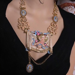 Peut inclure: Collier dor&eacute; orn&eacute; d'un pendentif central repr&eacute;sentant un ch&eacute;rubin, entour&eacute; de motifs floraux et de volutes. Le collier comprend des accents de style cam&eacute;e et un pendentif pendant, cr&eacute;ant une esth&eacute;tique vintage.