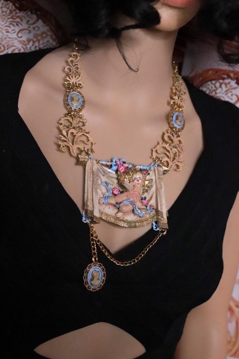 Peut inclure: Collier dor&eacute; orn&eacute; d'un pendentif central repr&eacute;sentant un ch&eacute;rubin, entour&eacute; de motifs floraux et de volutes. Le collier comprend des accents de style cam&eacute;e et un pendentif pendant, cr&eacute;ant une esth&eacute;tique vintage.