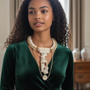 Puede incluir: Un collar llamativo con una banda blanca texturizada adornada con pequeñas perlas y detalles dorados. Un colgante central rectangular de estilo arquitectónico cuelga de la banda, conectado a una figura vertical tallada de color marfil y una cadena.
