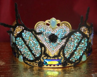 May include: A gold crown with black velvet and blue and clear rhinestones. The crown features two butterfly shapes and a heart shape in the center.