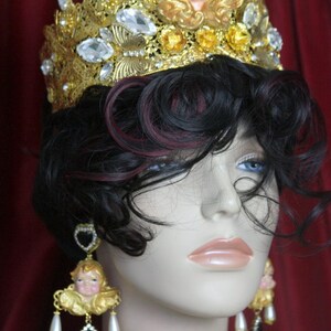 May include: A gold crown with a cherub figurine on top, adorned with rhinestones and pearls. The crown is worn by a mannequin with dark hair and a pair of dangling earrings featuring cherub figurines and black stones.