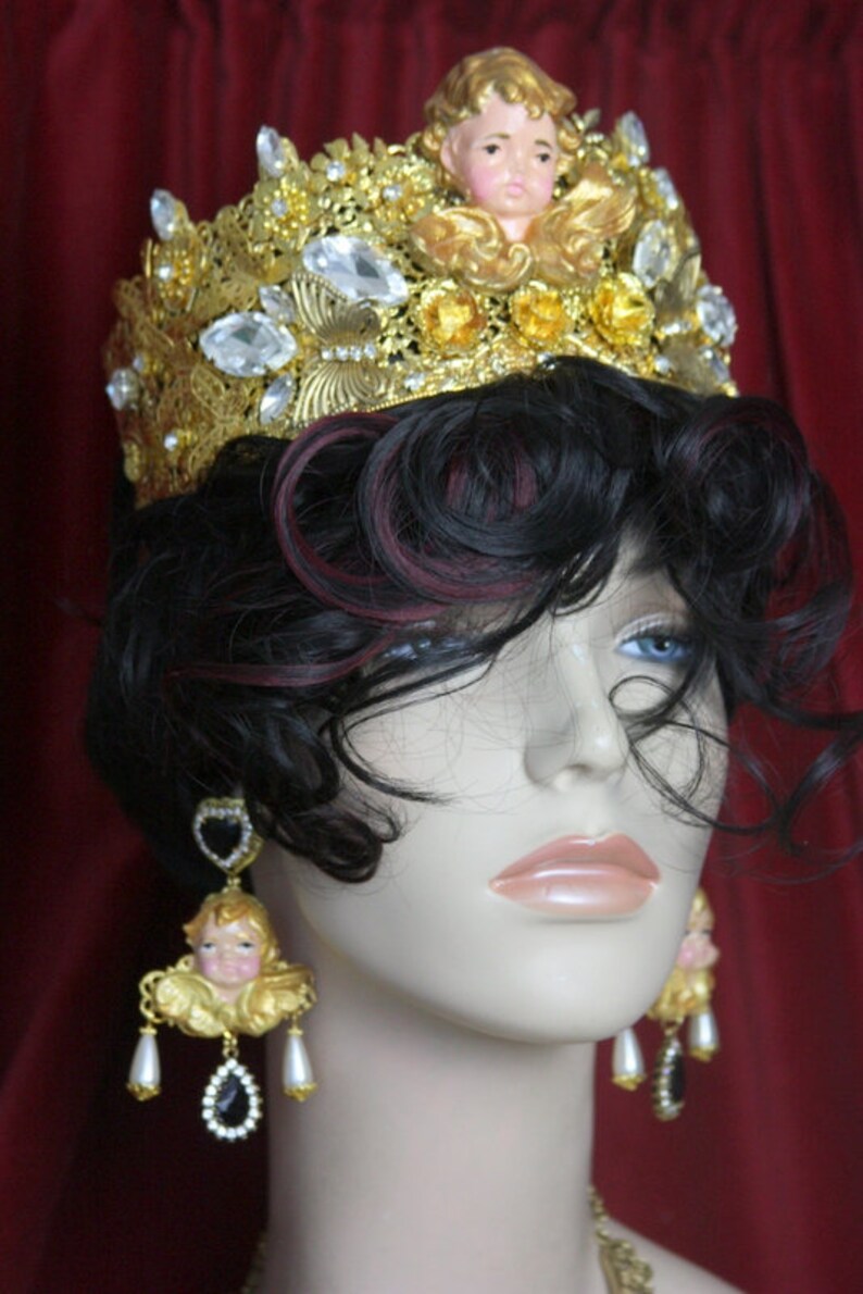 May include: A gold crown with a cherub figurine on top, adorned with rhinestones and pearls. The crown is worn by a mannequin with dark hair and a pair of dangling earrings featuring cherub figurines and black stones.