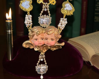 May include: A gold-toned necklace featuring cherub pendants, pearl accents, and ornate detailing. The necklace includes a central pendant with two cherub faces and smaller rectangular pendants. The design is set against a dark red velvet surface.