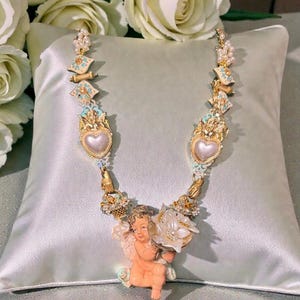 May include: A detailed necklace featuring a cherub pendant holding a flower. The necklace has gold-colored accents, pearls, and heart-shaped pendants. The design incorporates light blue and white enamel details, creating a vintage aesthetic.
