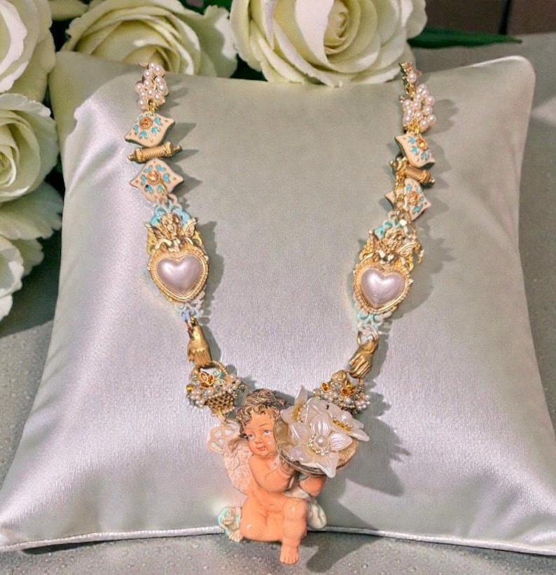 May include: A detailed necklace featuring a cherub pendant holding a flower. The necklace has gold-colored accents, pearls, and heart-shaped pendants. The design incorporates light blue and white enamel details, creating a vintage aesthetic.