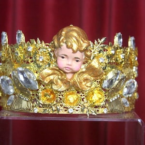 May include: A gold crown with a cherub figurine in the center. The crown is decorated with rhinestones and has a floral design.