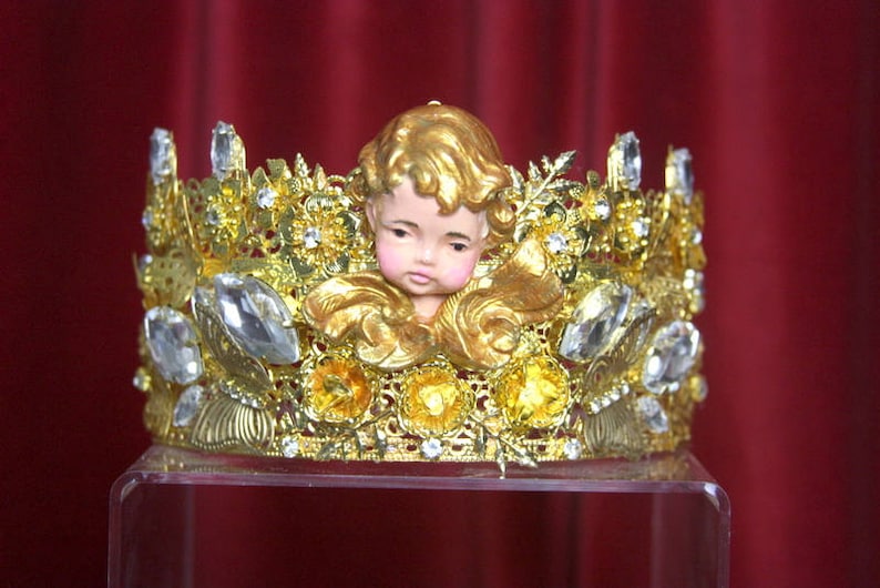 May include: A gold crown with a cherub figurine in the center. The crown is decorated with rhinestones and has a floral design.