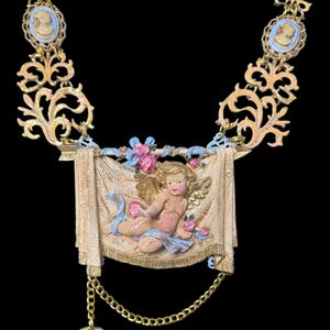 Peut inclure: Collier dor&eacute; orn&eacute; d'un ch&eacute;rubin sur une banni&egrave;re, agr&eacute;ment&eacute; de roses roses et de rubans bleus. Le collier comprend des pendentifs cam&eacute;e et un charme pendant, le tout sur fond noir. Une pi&egrave;ce de d&eacute;claration d'inspiration vintage.