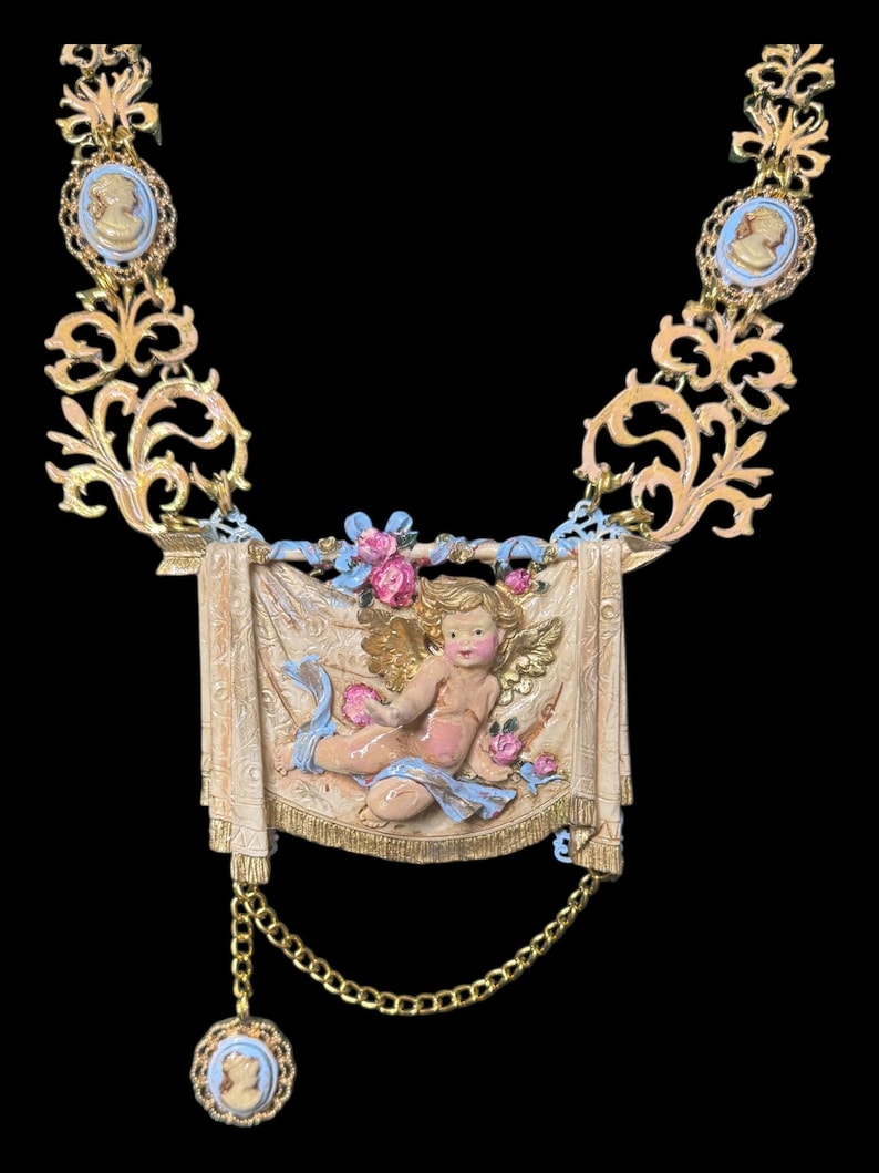 Peut inclure: Collier dor&eacute; orn&eacute; d'un ch&eacute;rubin sur une banni&egrave;re, agr&eacute;ment&eacute; de roses roses et de rubans bleus. Le collier comprend des pendentifs cam&eacute;e et un charme pendant, le tout sur fond noir. Une pi&egrave;ce de d&eacute;claration d'inspiration vintage.